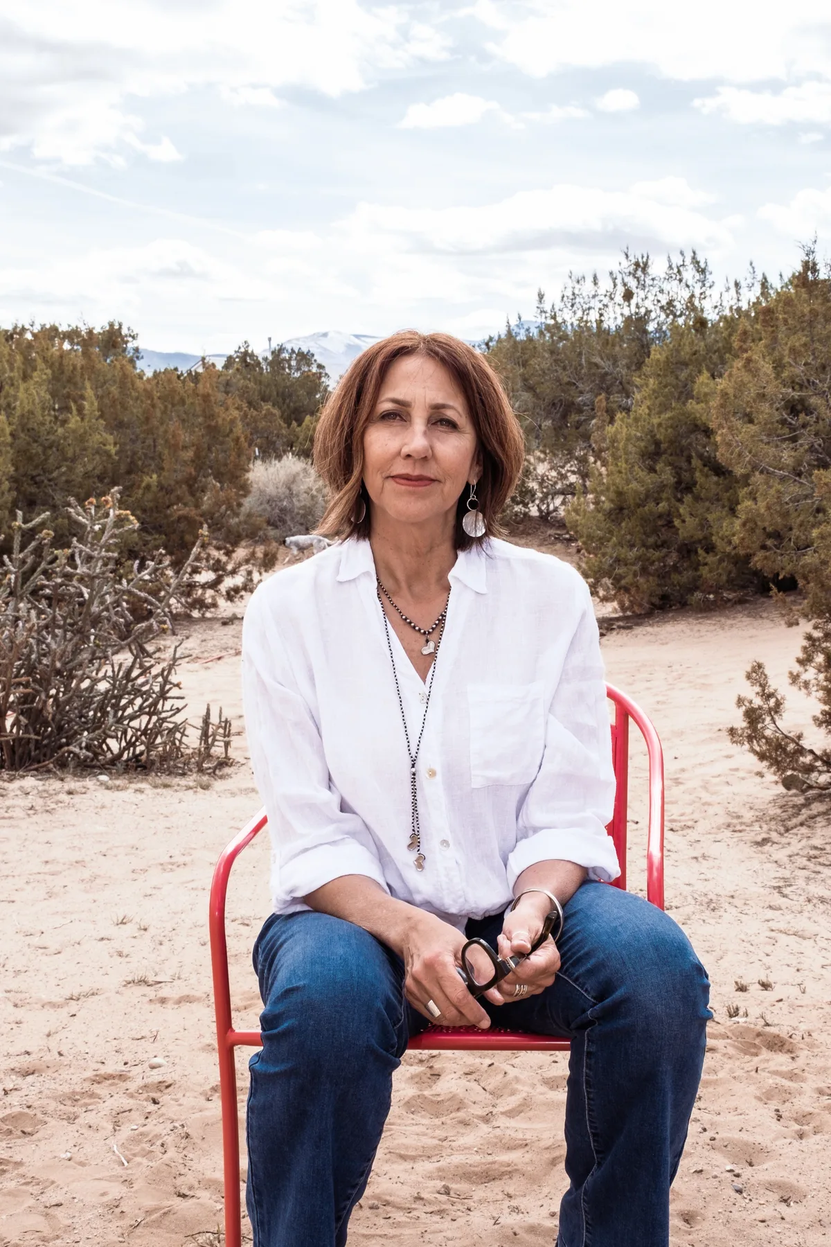 Linda Seebantz seated on a red chair in the high desert, juniper and mountains behind her